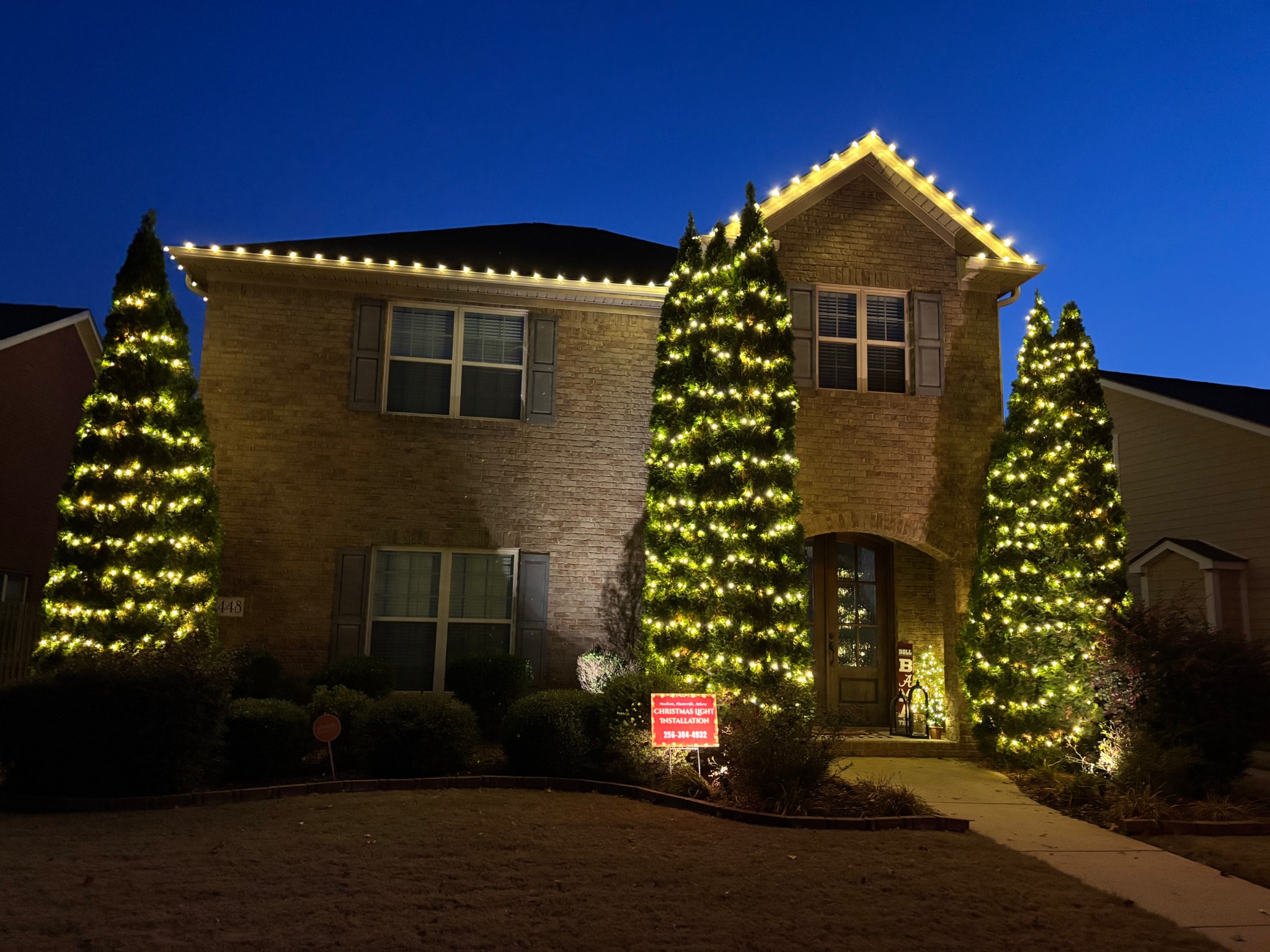 Warm white roofline and illuminated evergreen trees framing a brick home at dusk